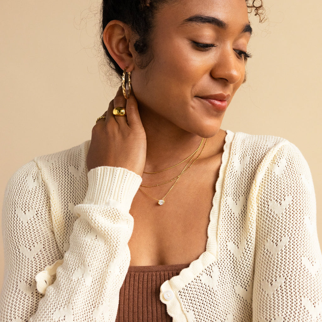 A woman smiles and looks down, her hand near her neck adorned with the Encased Hexagon Diamond Necklace, paired with a cream knit cardigan, brown top, and gold jewelry—a look of modern sophistication.