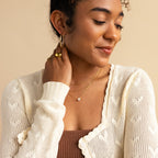A woman smiles and looks down, her hand near her neck adorned with the Encased Hexagon Diamond Necklace, paired with a cream knit cardigan, brown top, and gold jewelry—a look of modern sophistication.