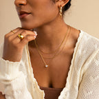 A woman wears layered gold necklaces, including the Encased Hexagon Diamond Necklace, along with gold rings and hoop earrings, styled with a cream knit cardigan over a brown top for a look of modern sophistication.