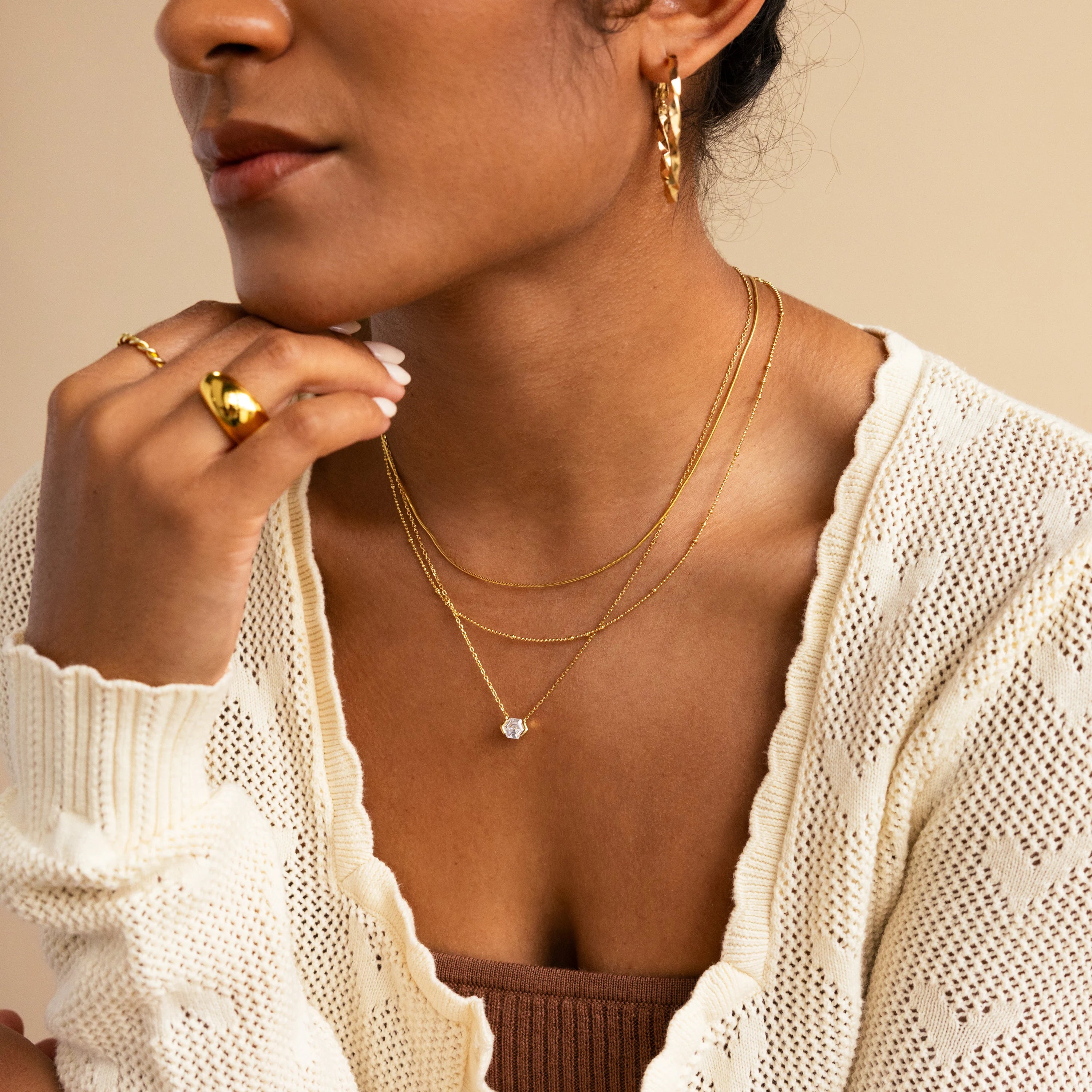 A woman wears layered gold necklaces, including the Encased Hexagon Diamond Necklace, along with gold rings and hoop earrings, styled with a cream knit cardigan over a brown top for a look of modern sophistication.