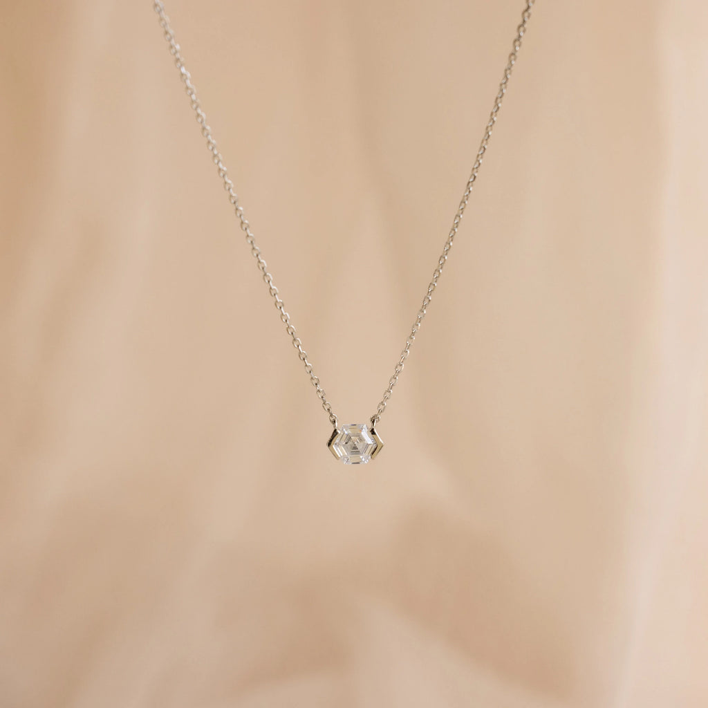 The Encased Hexagon Diamond Necklace features a clear diamond in a modern half-bezel setting on delicate silver, shown against a soft beige background.