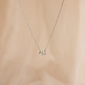 The Encased Hexagon Diamond Necklace features a clear diamond in a modern half-bezel setting on delicate silver, shown against a soft beige background.