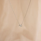 The Encased Hexagon Diamond Necklace features a clear diamond in a modern half-bezel setting on delicate silver, shown against a soft beige background.