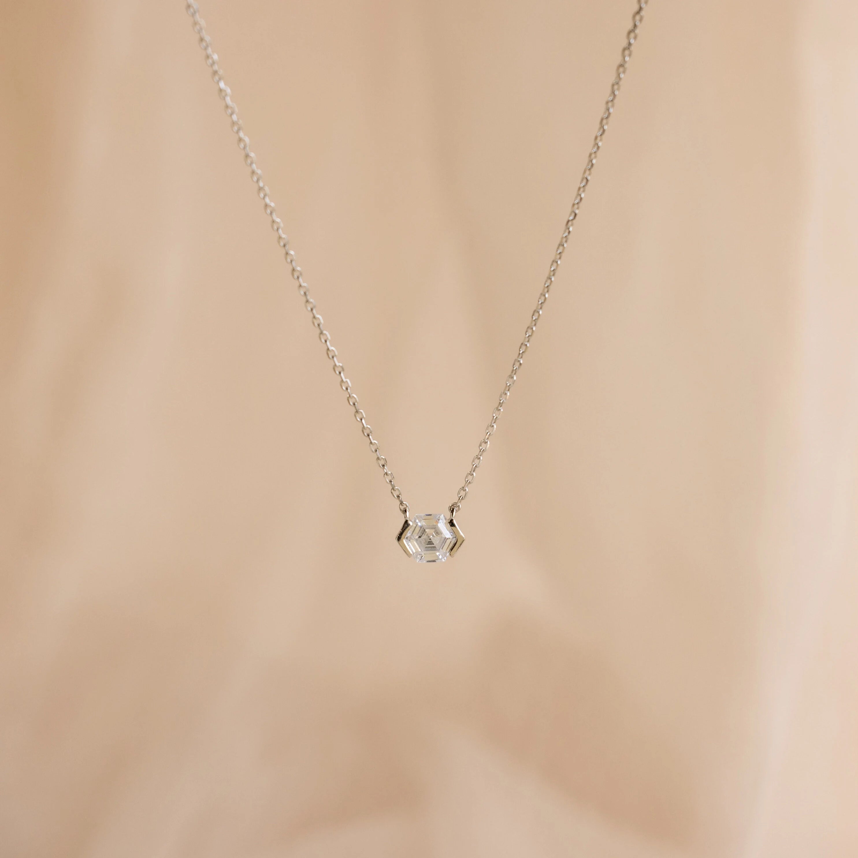 The Encased Hexagon Diamond Necklace features a clear diamond in a modern half-bezel setting on delicate silver, shown against a soft beige background.