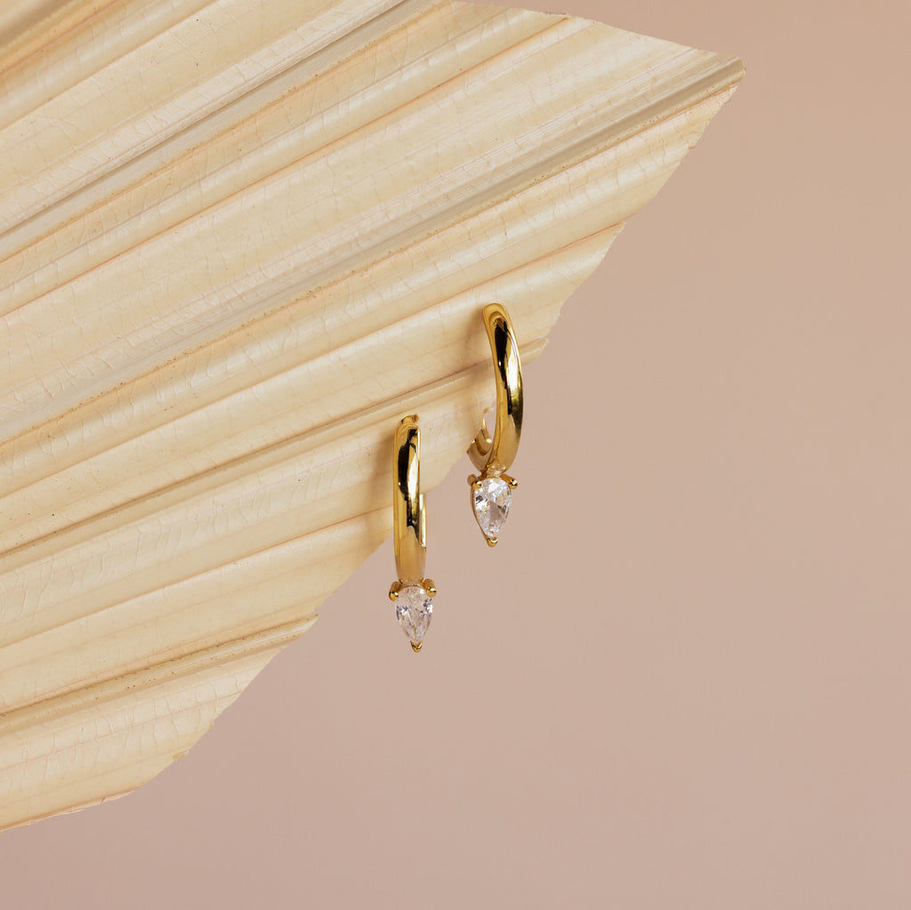 Elegant gold hoops with teardrop-shaped clear gemstones, the Pear Diamond Drop Hoops are a perfect wedding accessory, showcased on a textured cream-colored surface.