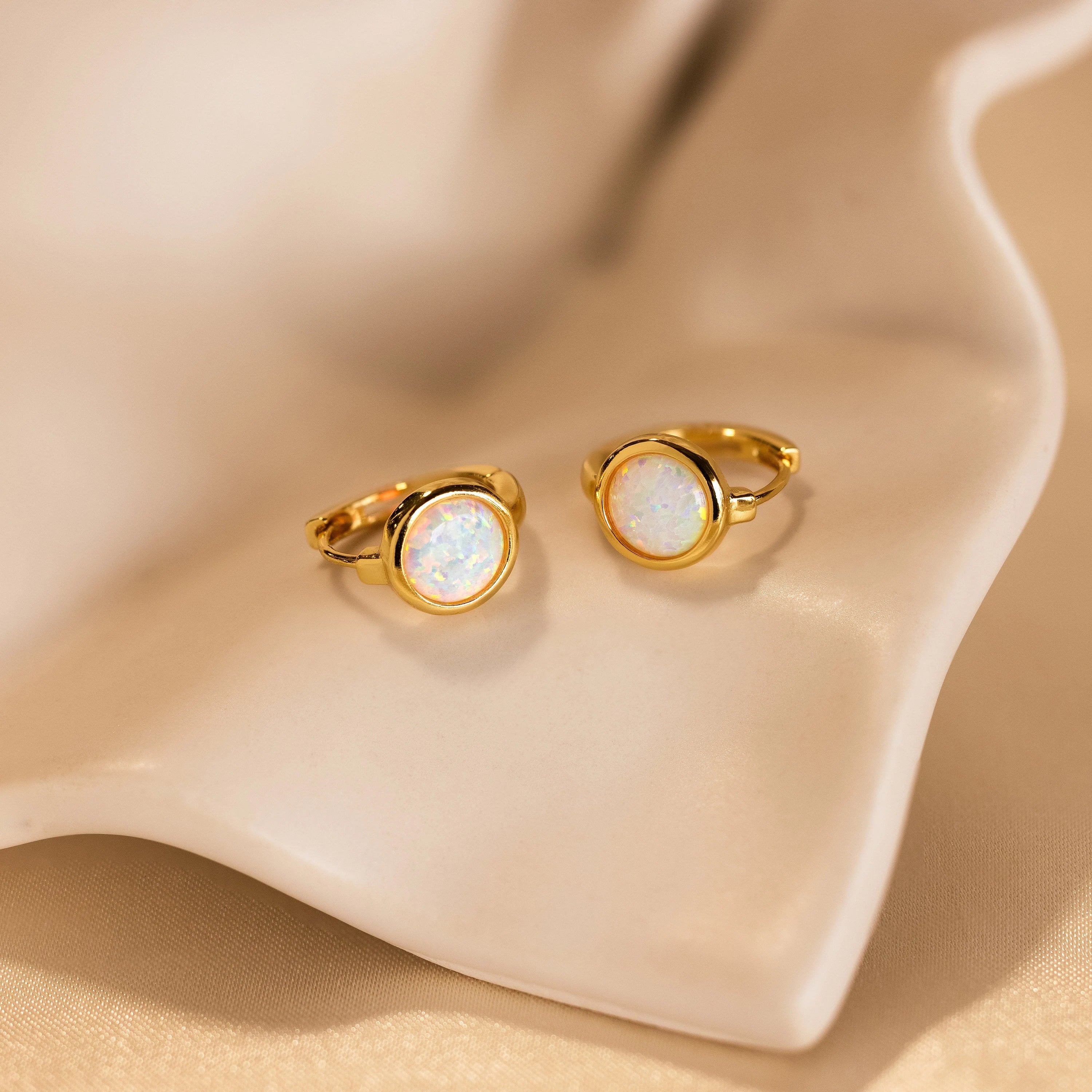 Displayed on a wavy, cream-colored surface, the Opal Signet Huggies are gold hoop earrings featuring dainty round opal stones.
