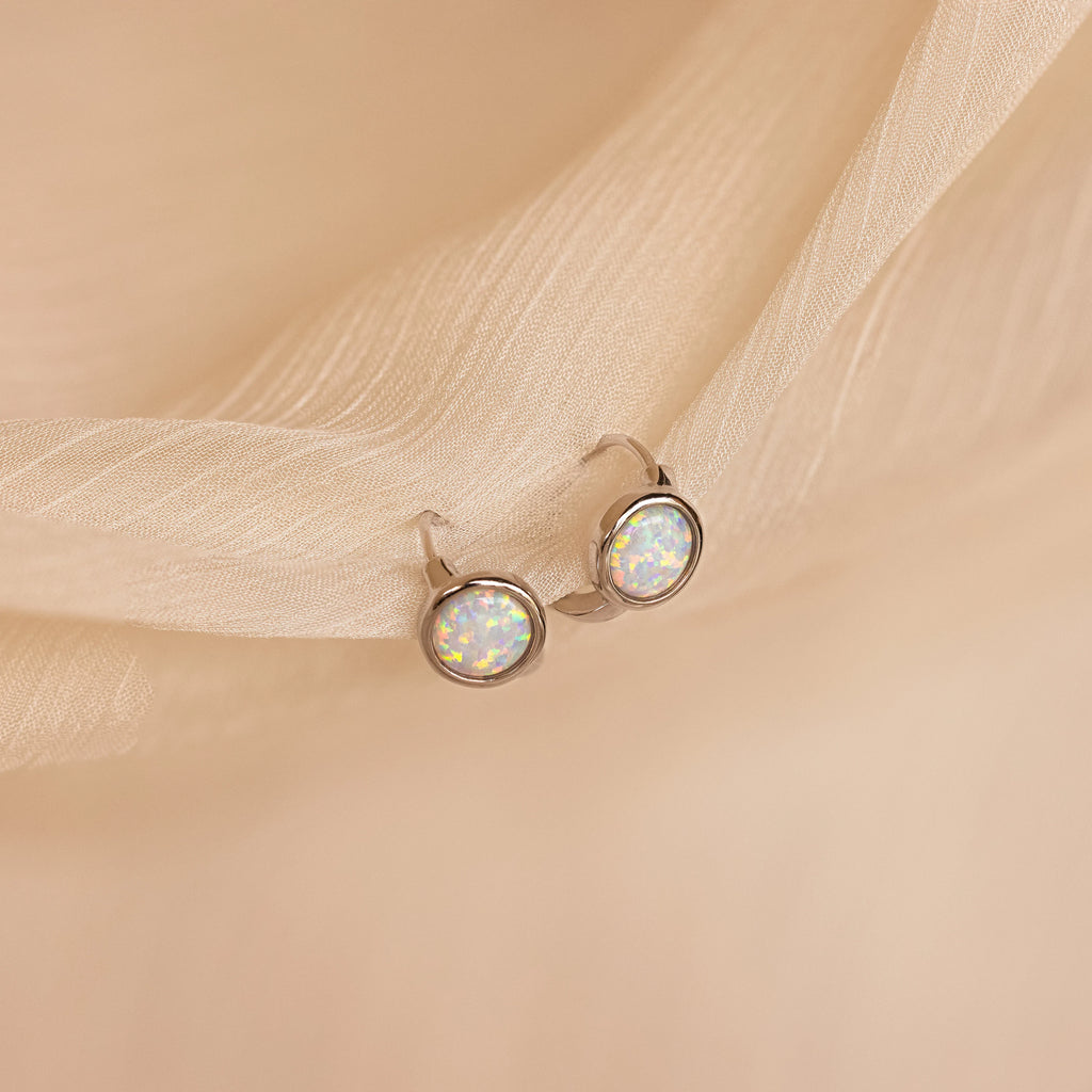 Opal Signet Huggies featuring round opal gemstones evoke the look of dainty hoops, shown against a soft, light beige fabric background.