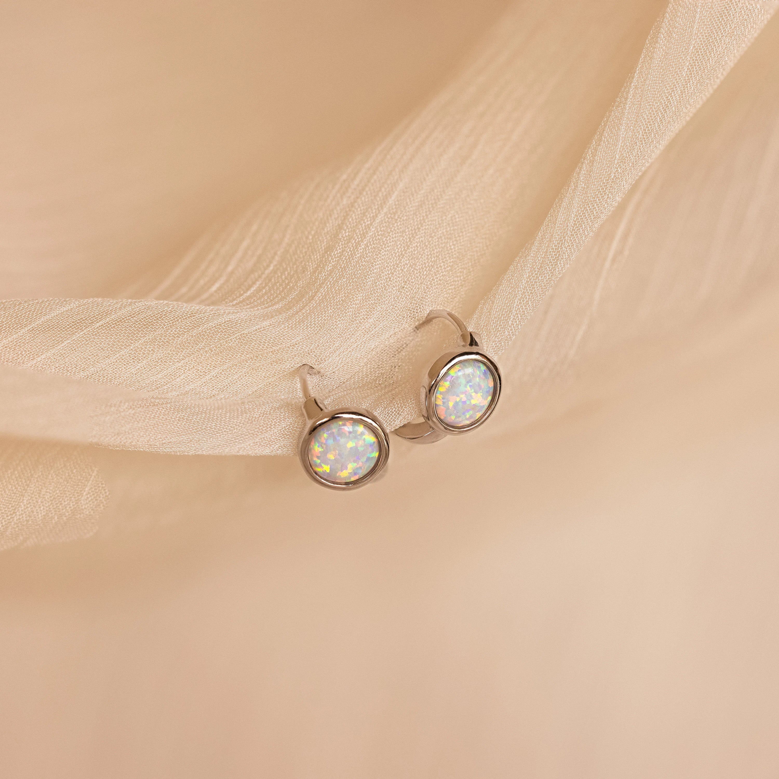 Opal Signet Huggies featuring round opal gemstones evoke the look of dainty hoops, shown against a soft, light beige fabric background.