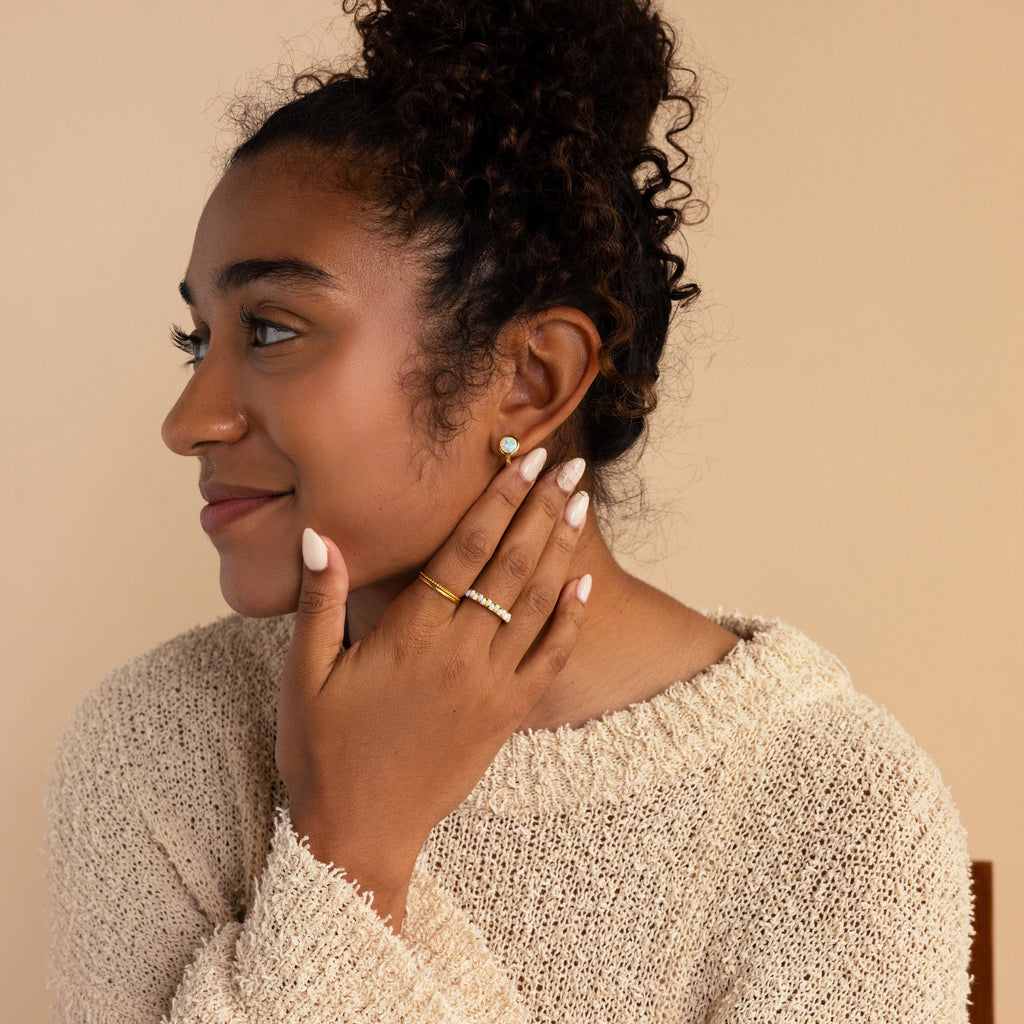 Woman with curly hair in a beige sweater, wearing Opal Signet Huggies earrings and gold rings, looking to the side.