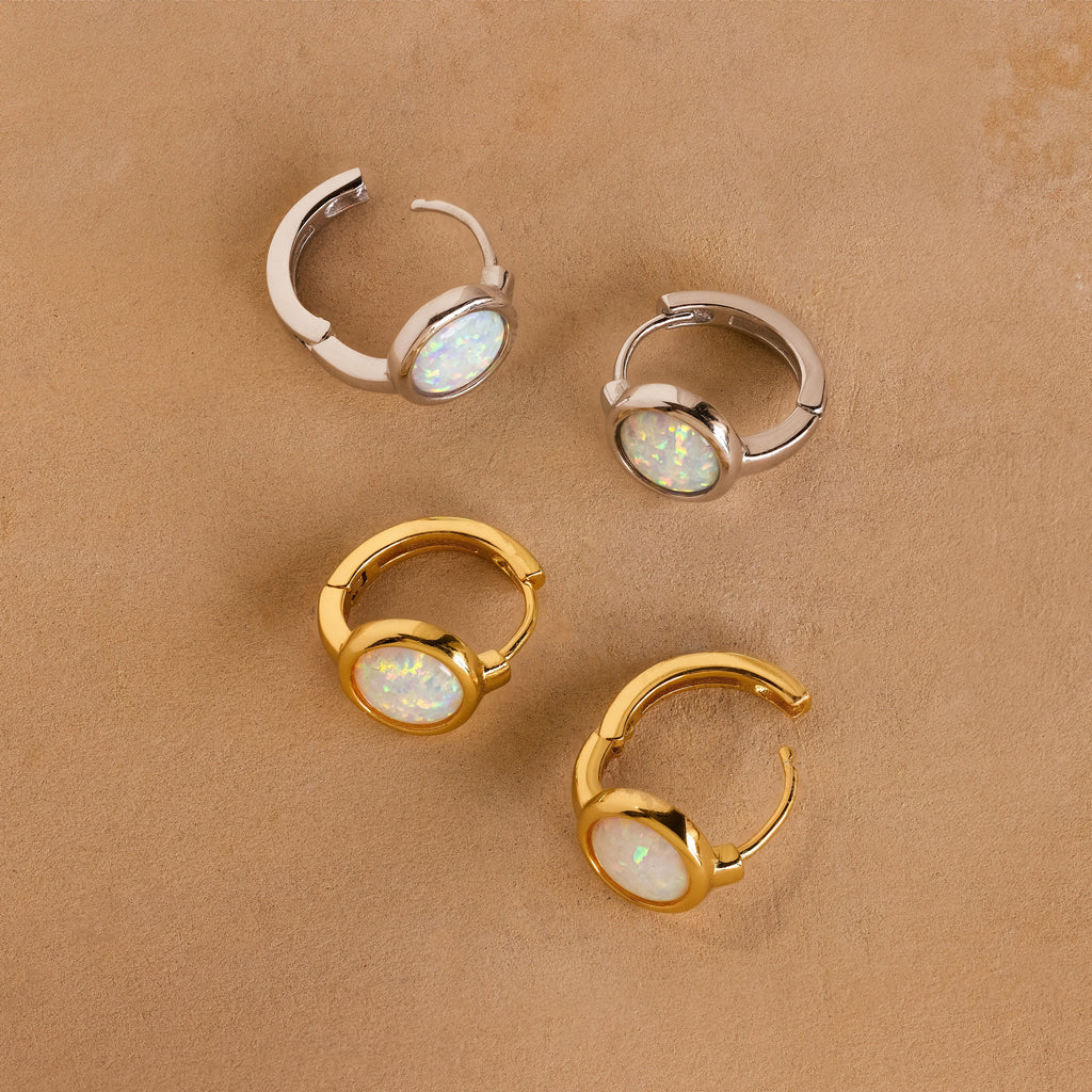 Four Opal Signet Huggies—two gold, two silver with round opals—are displayed on a tan textured surface, ideal for lovers of dainty hoops and understated style.