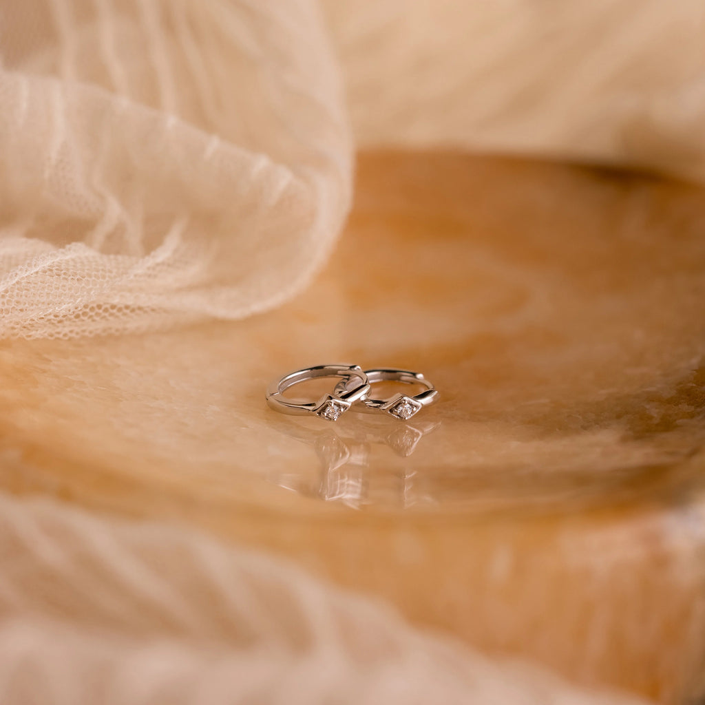 Two silver rings with small diamonds rest on marble, surrounded by beige fabric—the perfect complement to Ericka Diamond Hoops for an elegant, timeless look.