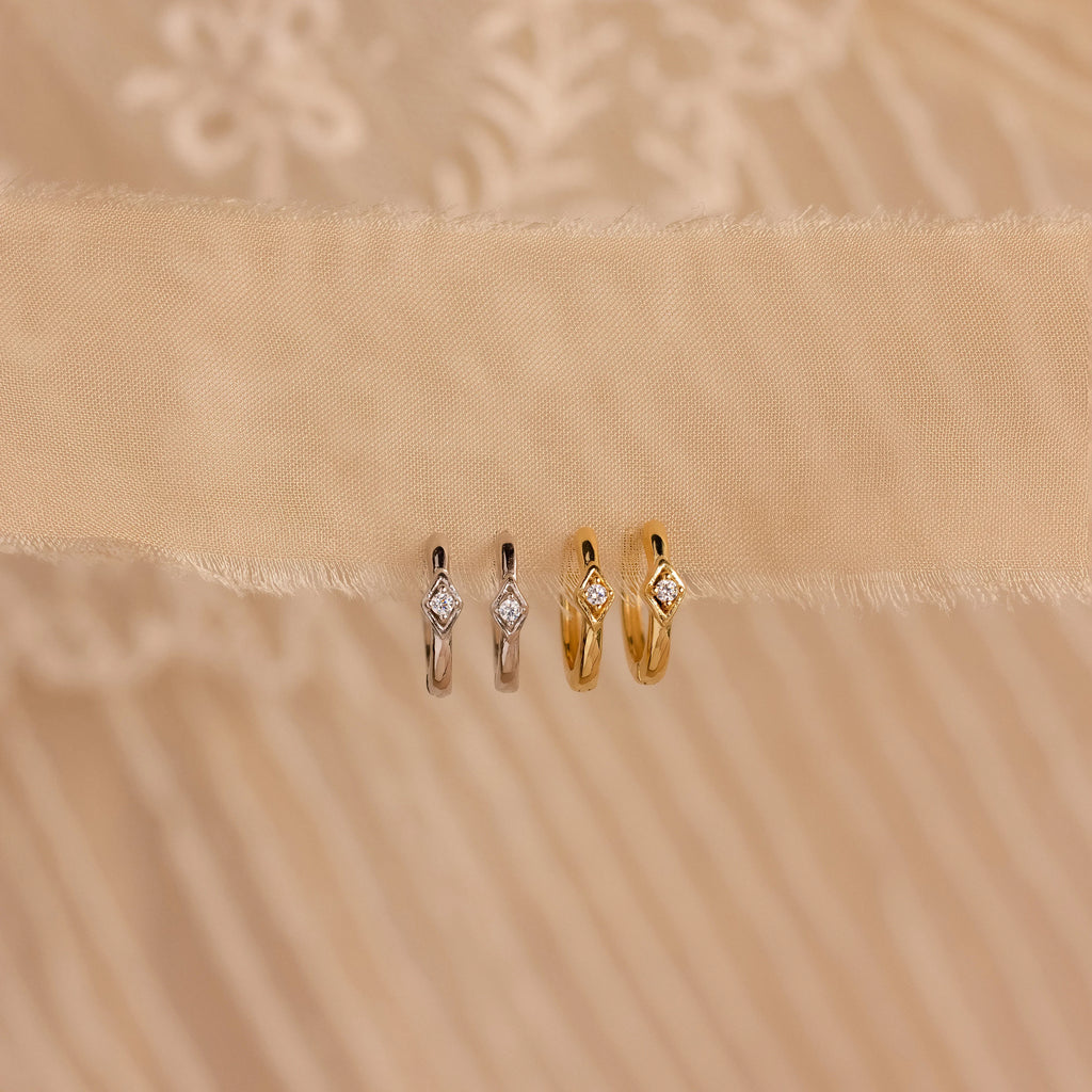 Two pairs of Ericka Diamond Hoops, one in silver and one in gold, each featuring a diamond, are displayed on beige fabric.