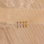 Two pairs of Ericka Diamond Hoops, one in silver and one in gold, each featuring a diamond, are displayed on beige fabric.