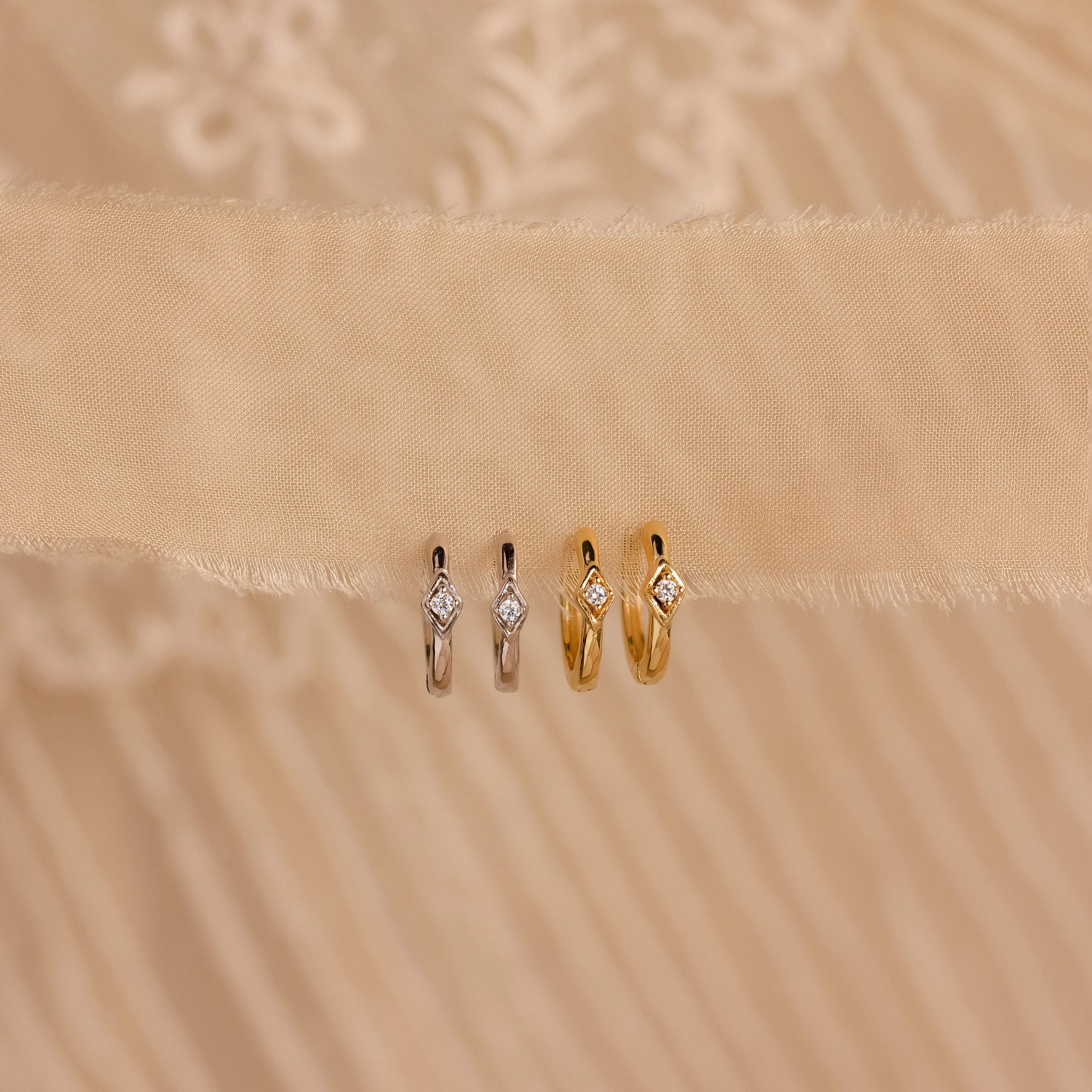 Two pairs of Ericka Diamond Hoops, one in silver and one in gold, each featuring a diamond, are displayed on beige fabric.