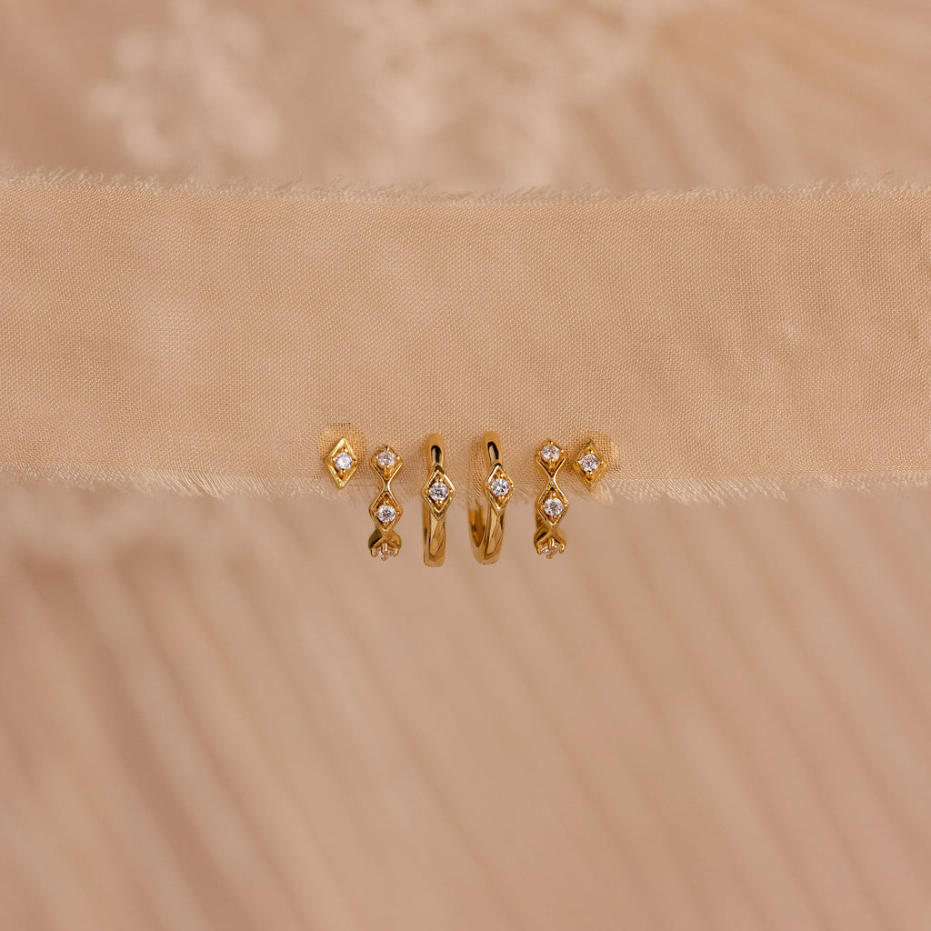 Five pairs of gold Ericka Diamond Hoops and other minimalist huggie earrings, featuring small diamonds, are displayed on a beige textured fabric background.