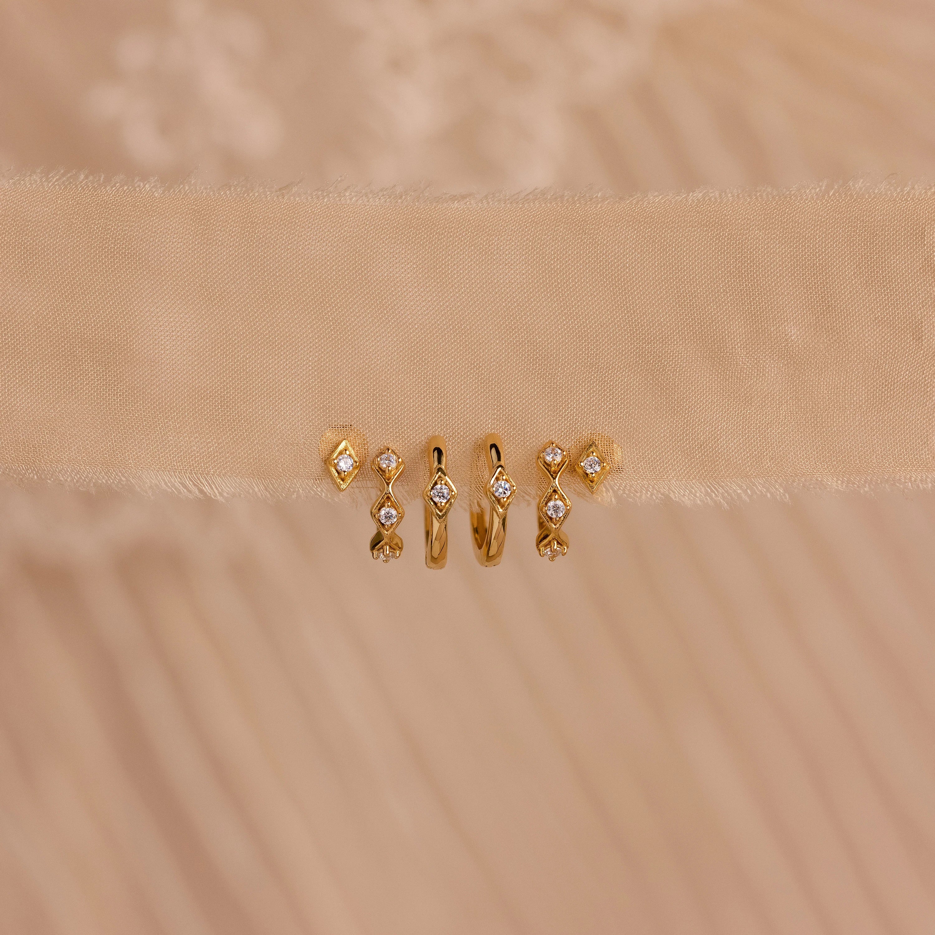 Five pairs of gold Ericka Diamond Hoops and other minimalist huggie earrings, featuring small diamonds, are displayed on a beige textured fabric background.