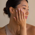 A woman with manicured nails and a Diamond Station Ring gently touches her face, gazing sideways with a neutral expression.