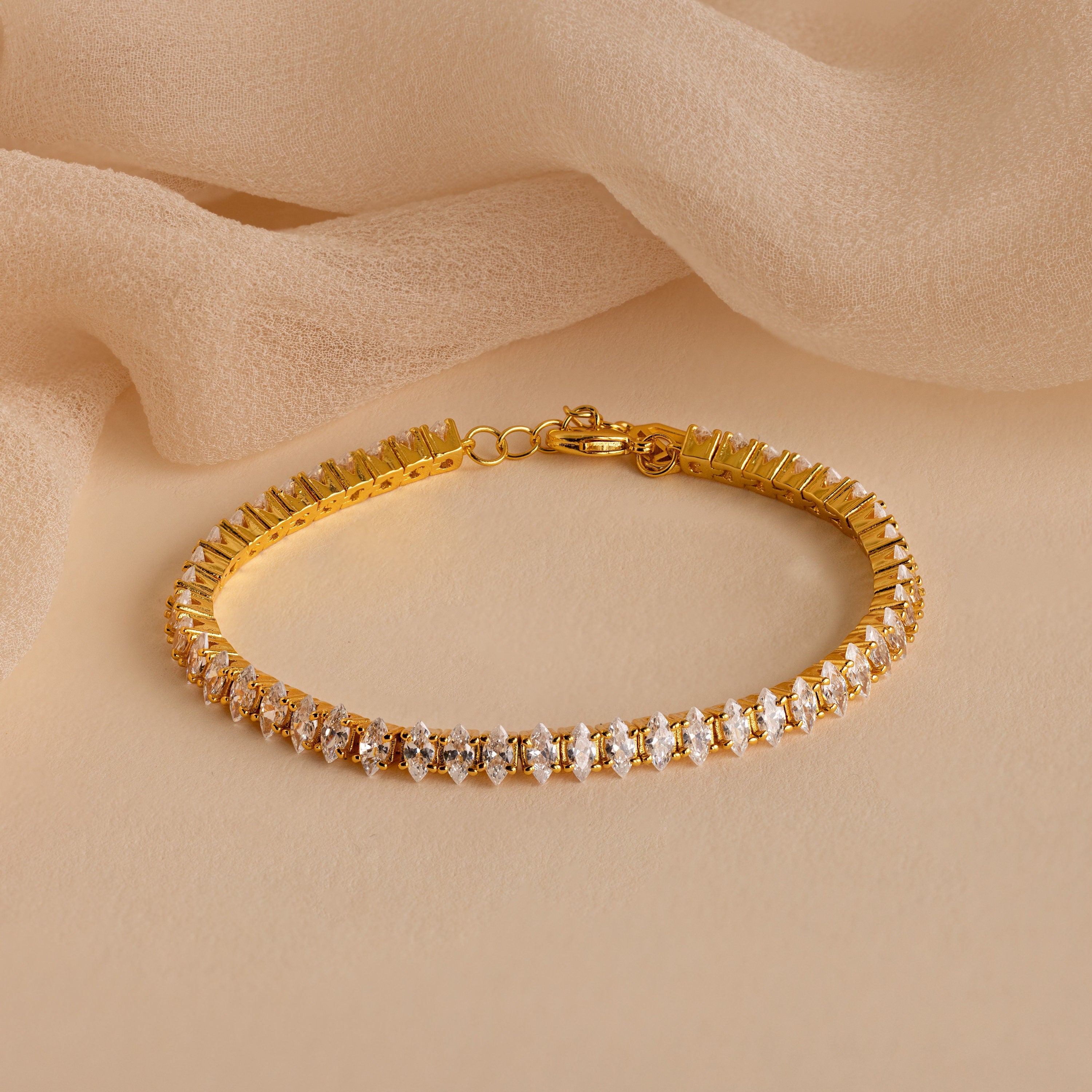 The Marquise Diamond Tennis Bracelet displayed flat on a beige fabric background, highlighting its sparkling marquise-cut stones and polished gold setting.