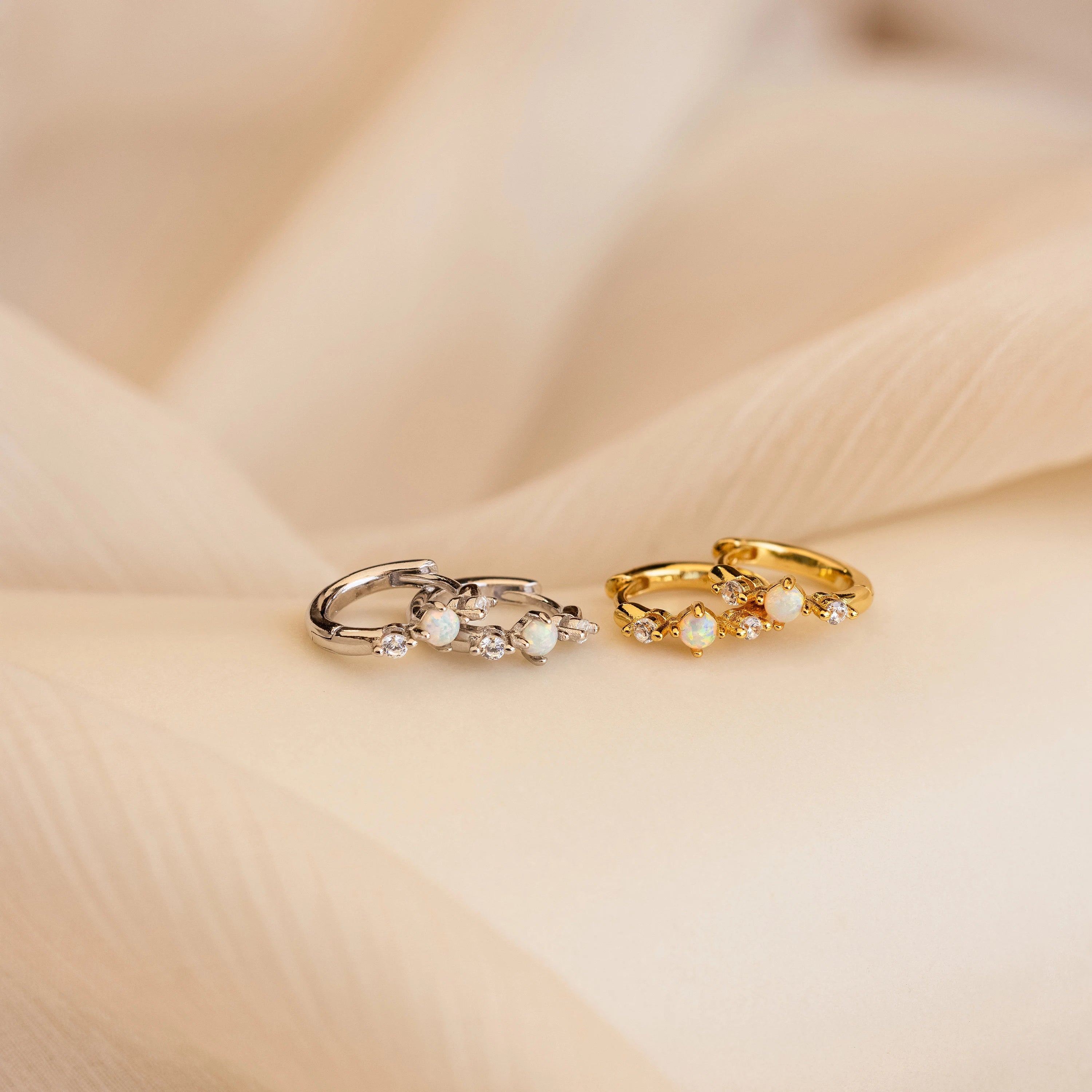 A pair of gold and a pair of silver huggie earrings resting on soft beige fabric, each adorned with a central opal gemstone and two small diamonds on either side, showcasing both metal color options side by side.