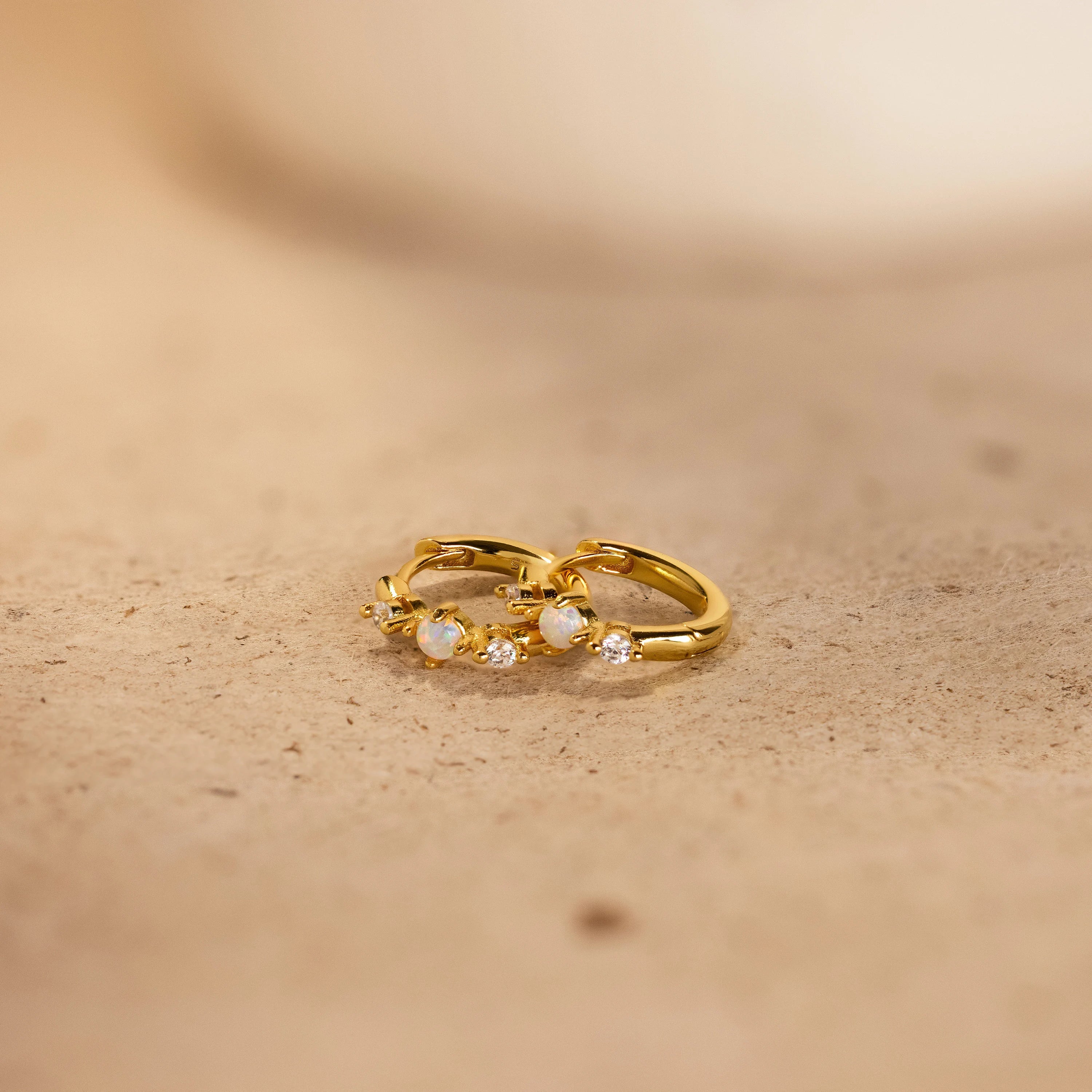 A pair of gold Opal Diamond Huggies laying on top of each other. The design has an opal gem in the middle with a diamond stone on each side. 