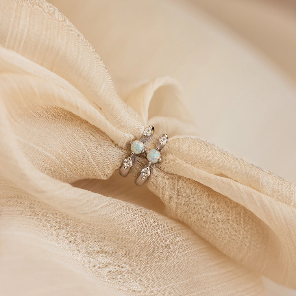Two silver opal and diamond huggie earrings styled together on softly gathered beige chiffon fabric, emphasizing the delicate shimmer and lustrous opal glow.