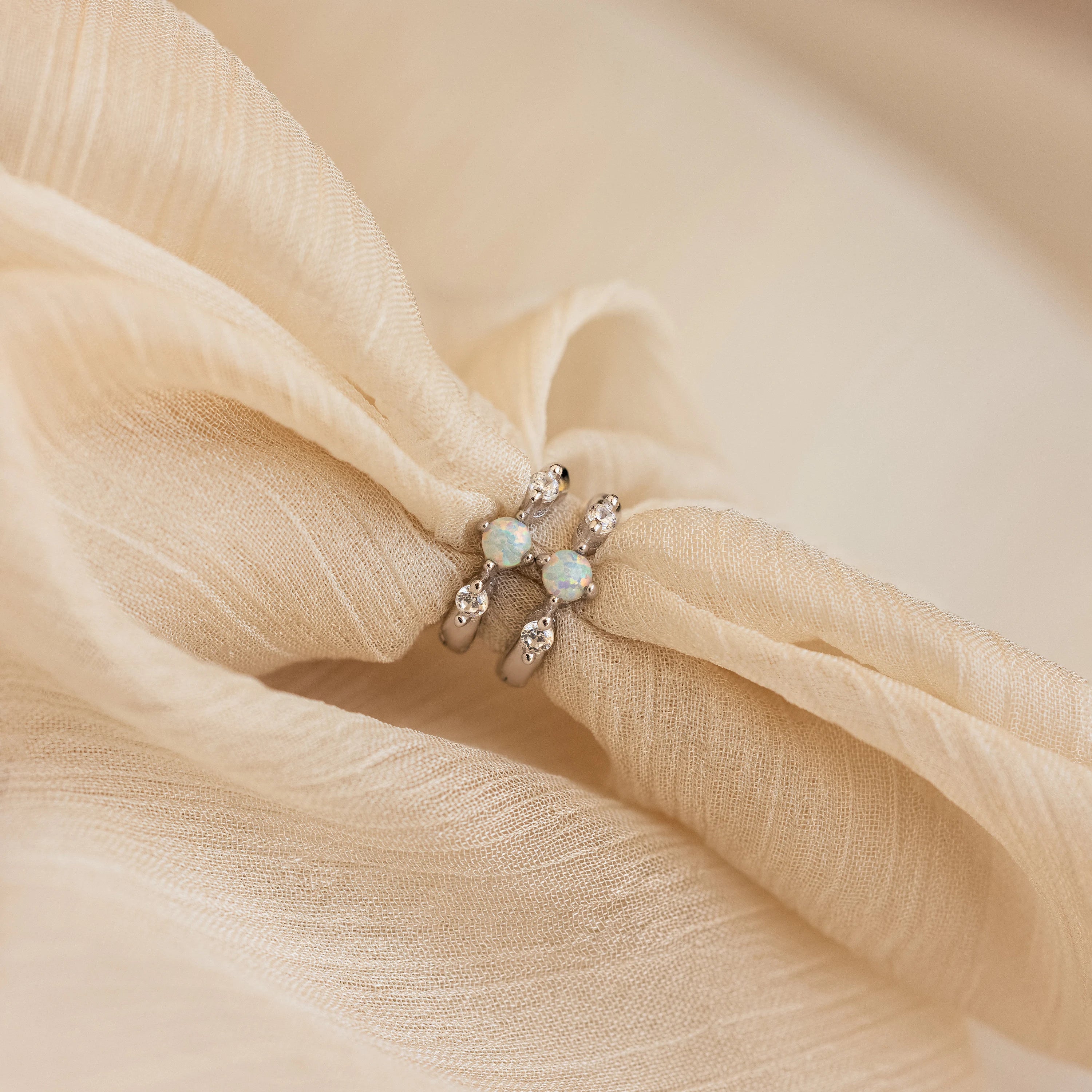 Two silver opal and diamond huggie earrings styled together on softly gathered beige chiffon fabric, emphasizing the delicate shimmer and lustrous opal glow.