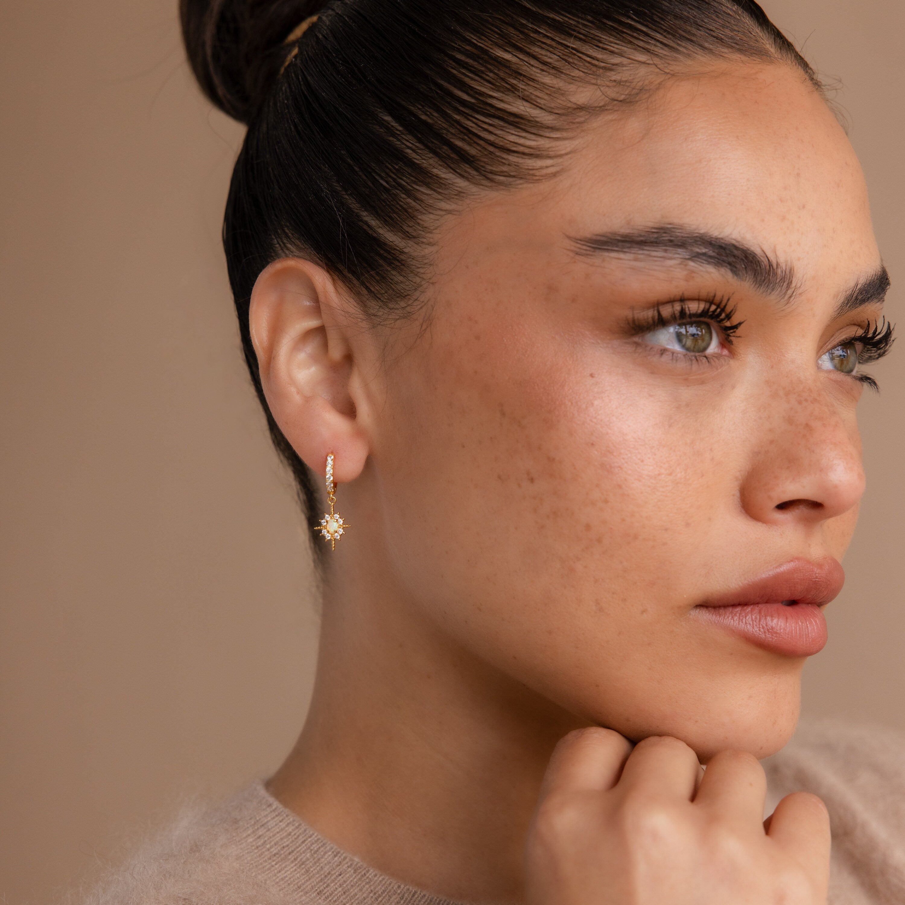 Woman with brown hair in a bun wearing Pave Opal Star Huggies, looking thoughtfully to the side.