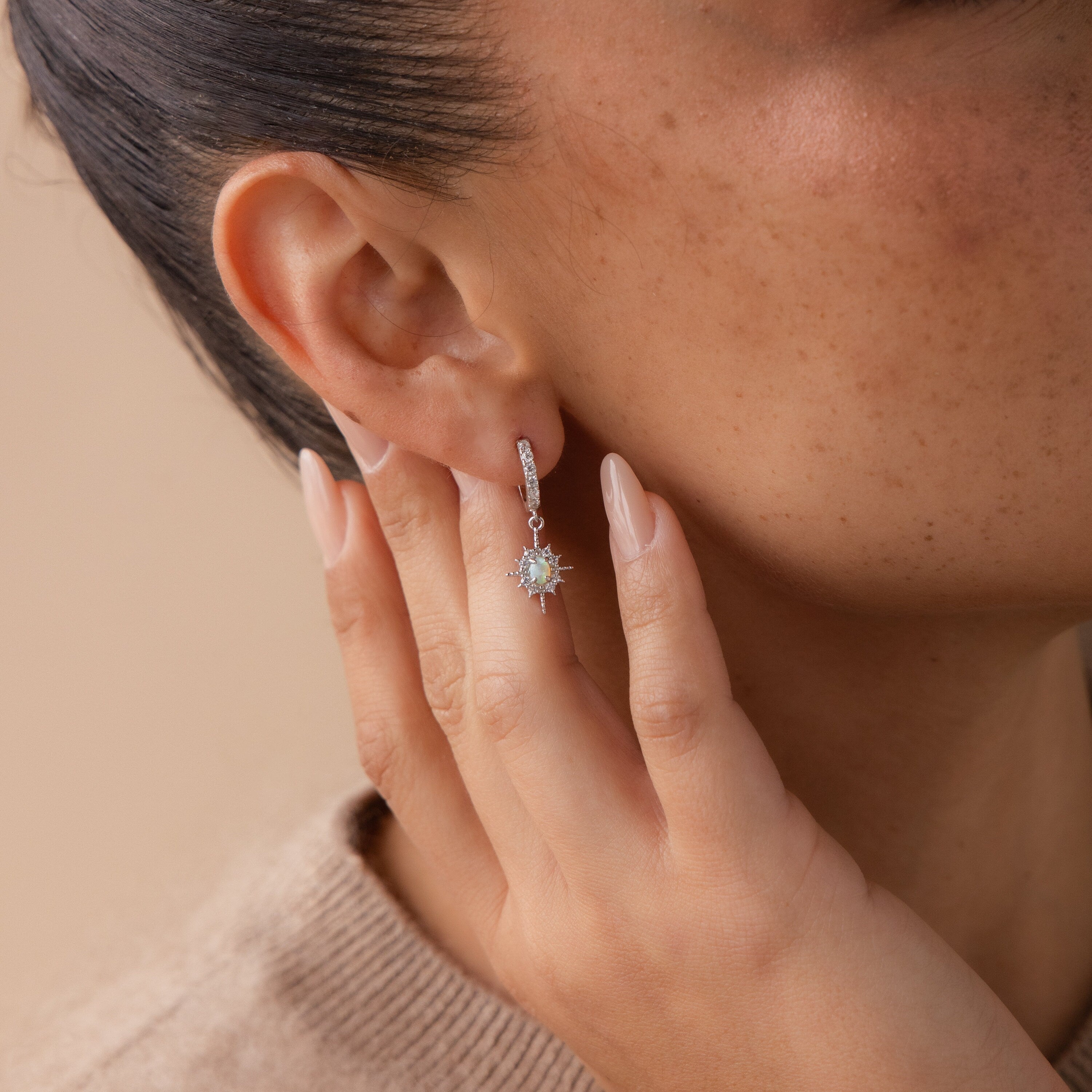 A woman touches her ear, showing off the Pave Opal Star Huggies—delicate hoop earrings adorned with a sparkling light blue opal star charm.
