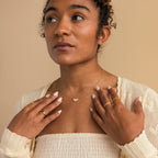 A woman in a cream top and gold jewelry, featuring the White Opal Marquise Necklace, looks up with her hands near her collarbone against a beige background.