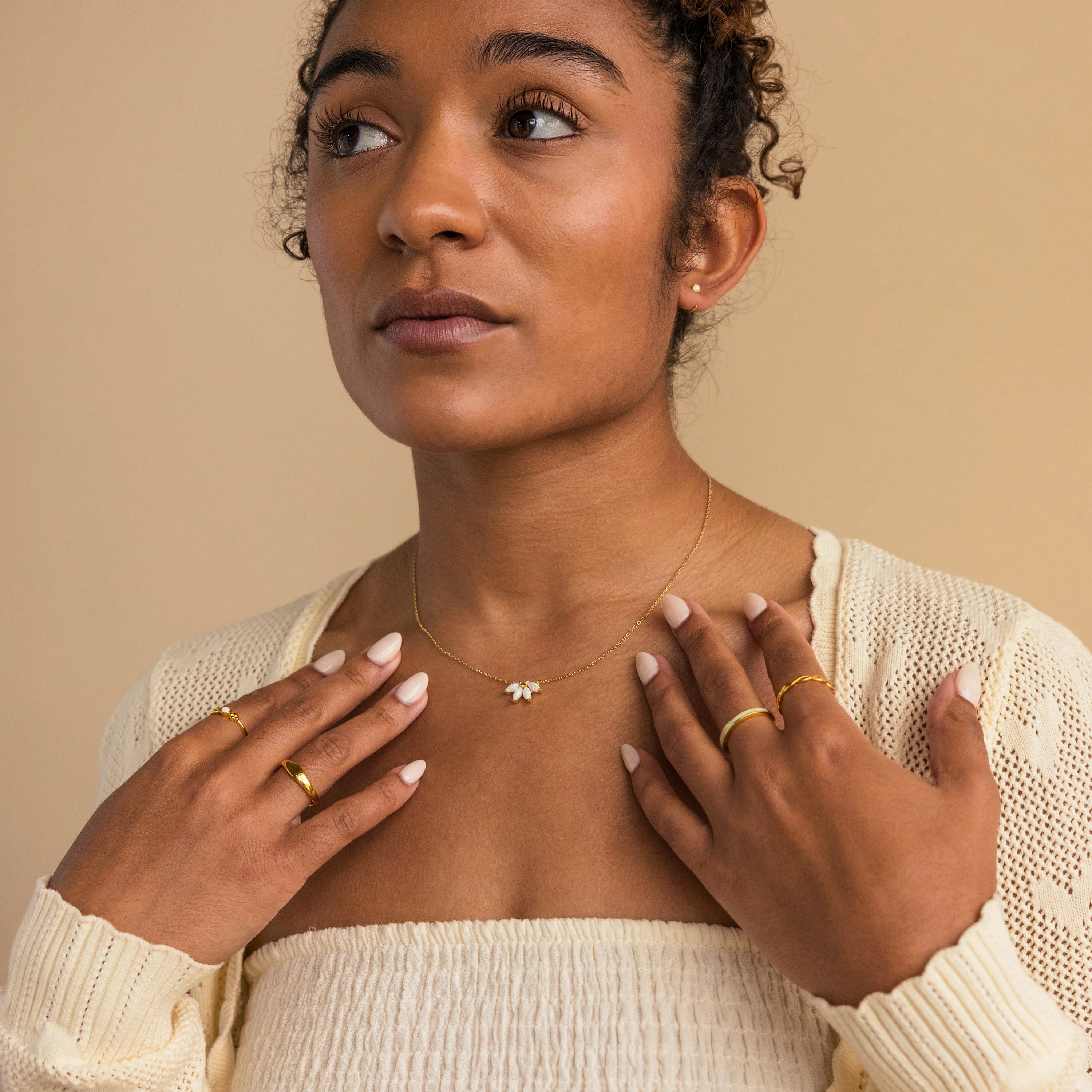A woman in a cream top and gold jewelry, featuring the White Opal Marquise Necklace, looks up with her hands near her collarbone against a beige background.
