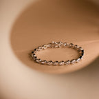 The Pear Diamond Tennis Bracelet, featuring dazzling oval gemstones, is showcased on a beige background framed by a gentle circular blur.