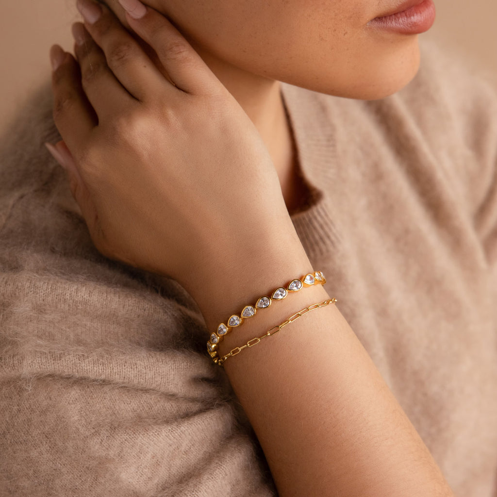Close-up of a woman wearing two gold bracelets, featuring the Pear Diamond Tennis Bracelet adorned with heart-shaped gems, as she rests her hand on her neck.