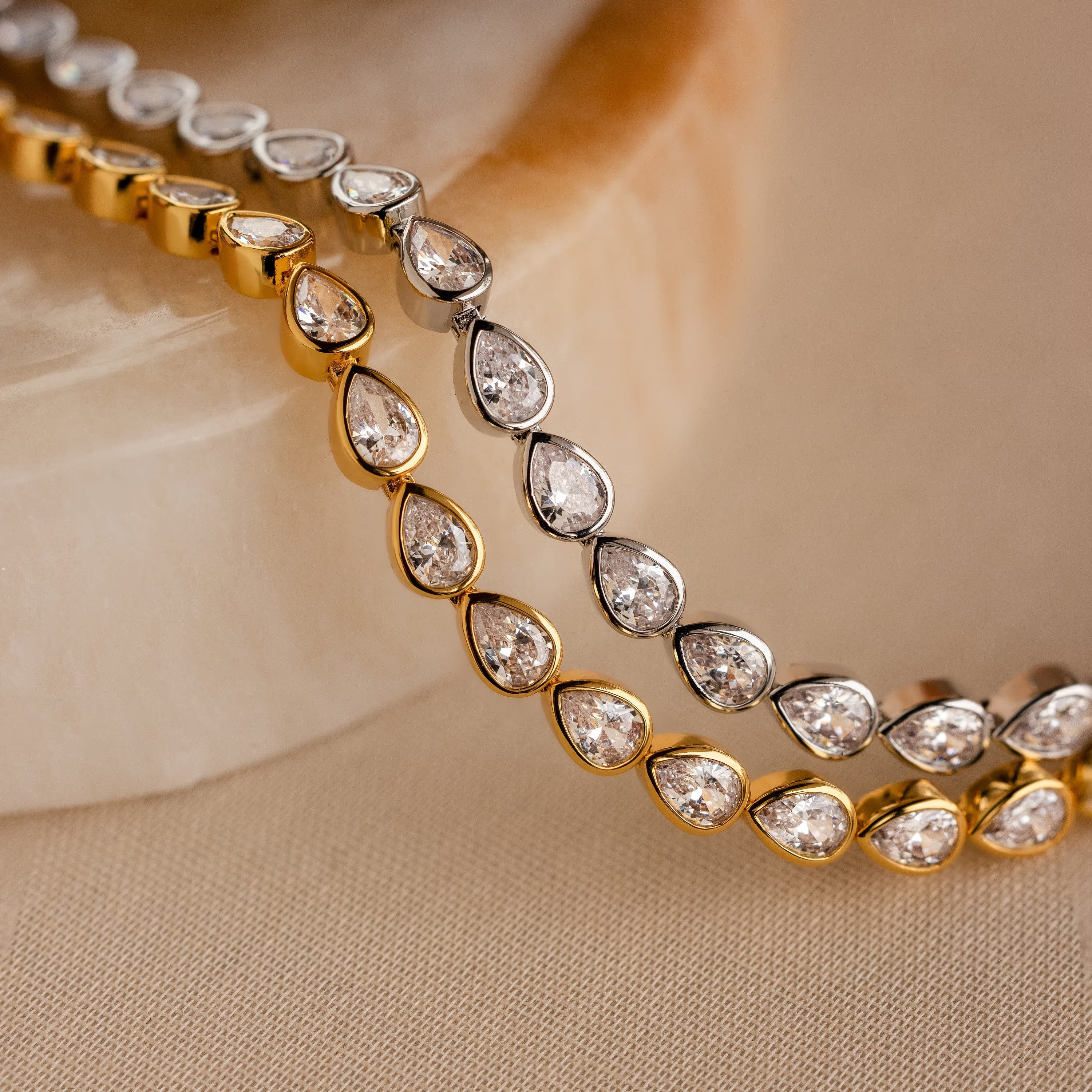 The Pear Diamond Tennis Bracelet is shown in both gold and silver settings on a beige surface, highlighting the timeless elegance of pear-cut diamonds.