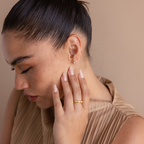 Woman with closed eyes touching her face, wearing the Pave Opal Ring Set—elegant gold bands with a pavé opal accent—paired with a pleated beige top against a neutral background.