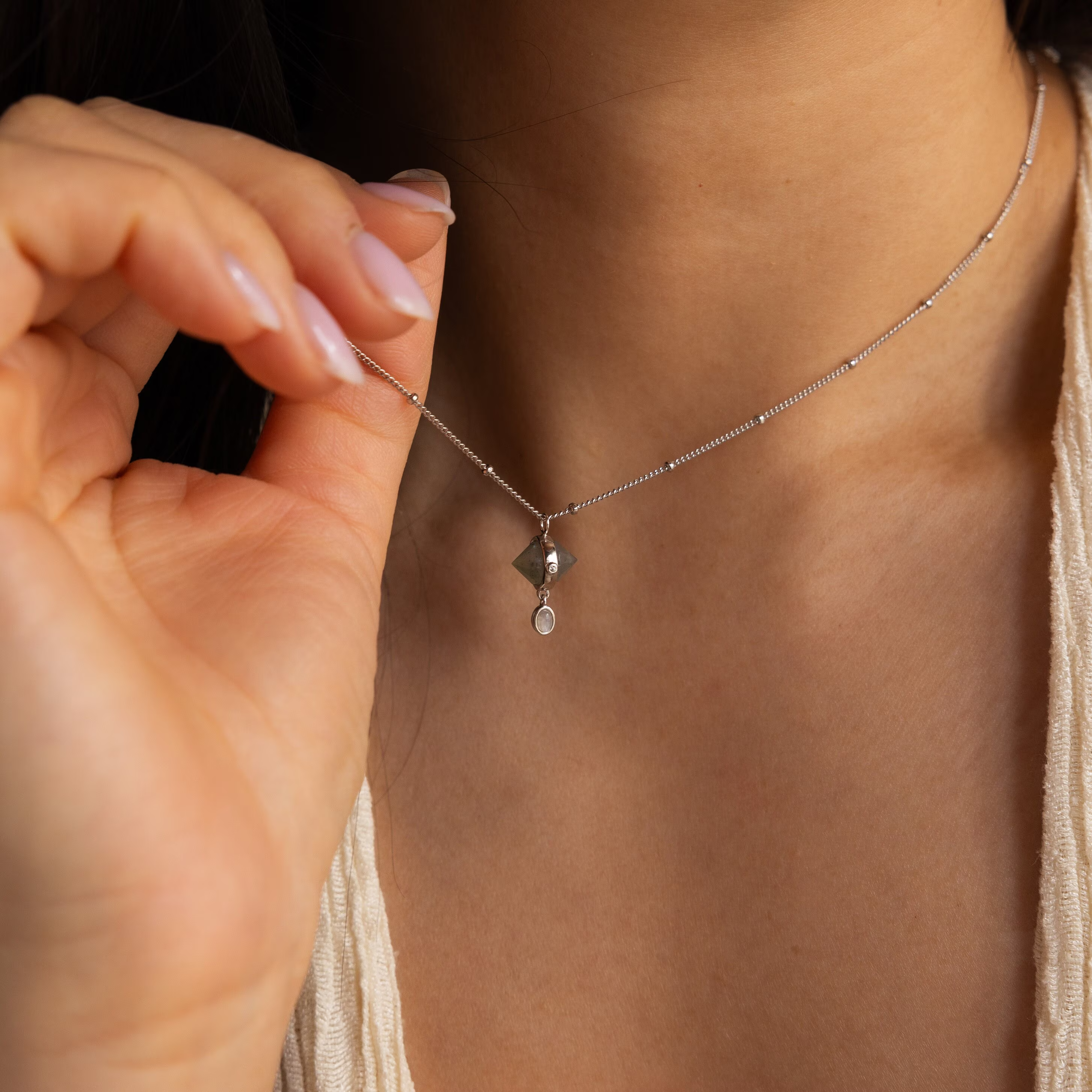 Close-up of a woman holding the Labradorite & Moonstone Drop Necklace—a delicate silver necklace with two small pendants—near her collarbone.