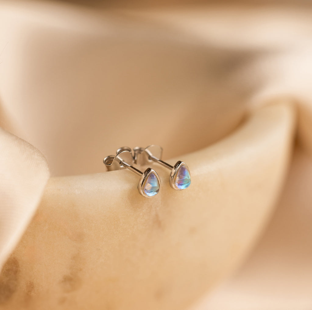 The Pear Moonstone Studs, set in silver and beautifully teardrop-shaped, add celestial charm to any look and are displayed on a smooth, beige surface.