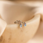 The Pear Moonstone Studs, set in silver and beautifully teardrop-shaped, add celestial charm to any look and are displayed on a smooth, beige surface.