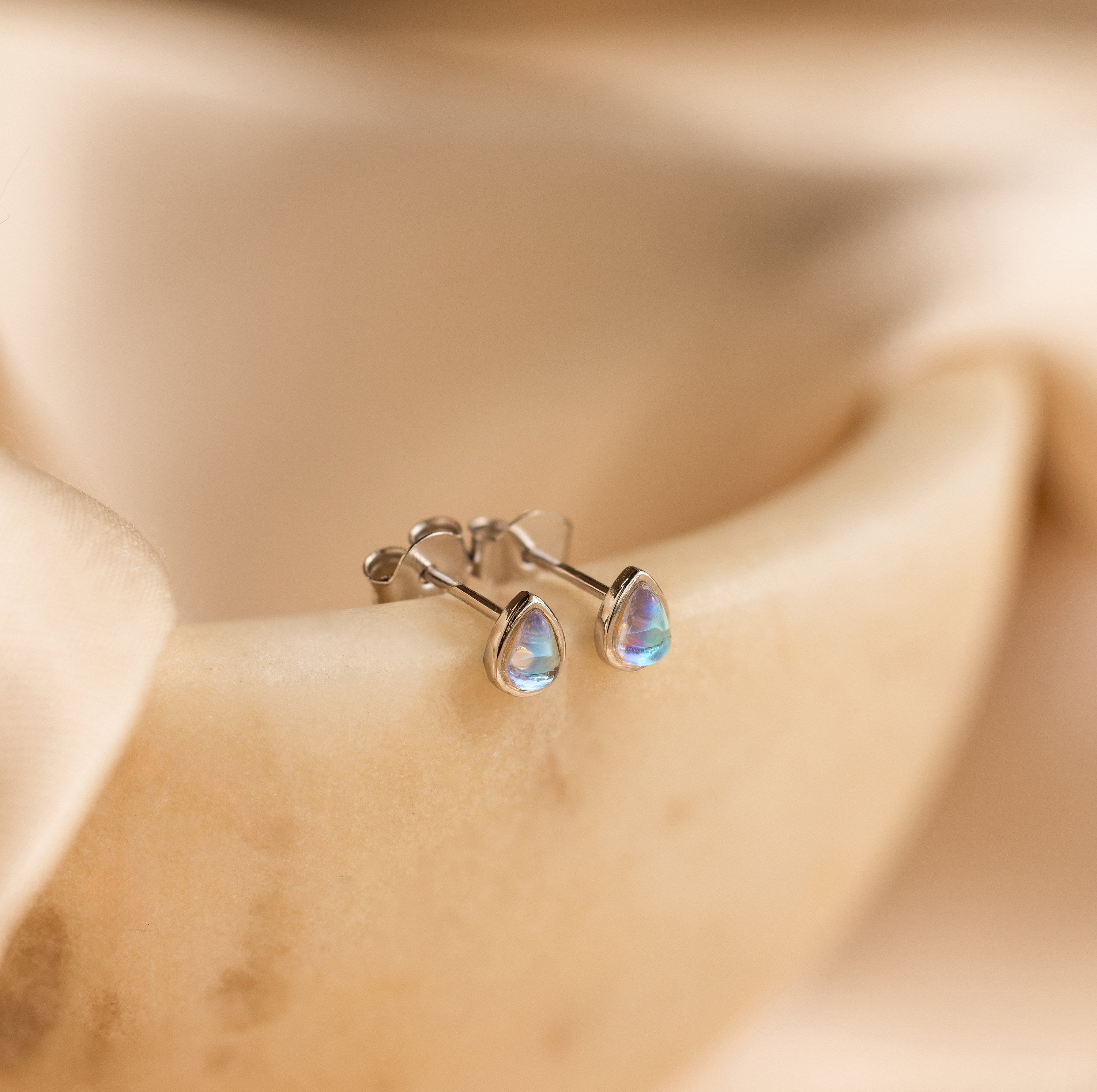 The Pear Moonstone Studs, set in silver and beautifully teardrop-shaped, add celestial charm to any look and are displayed on a smooth, beige surface.