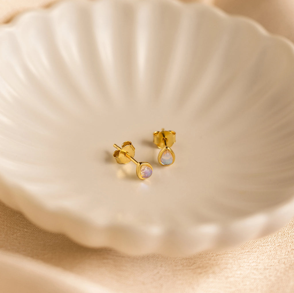 Pear Moonstone Studs with small iridescent stones rest on a white, scalloped dish—these dainty gold earrings radiate celestial charm.
