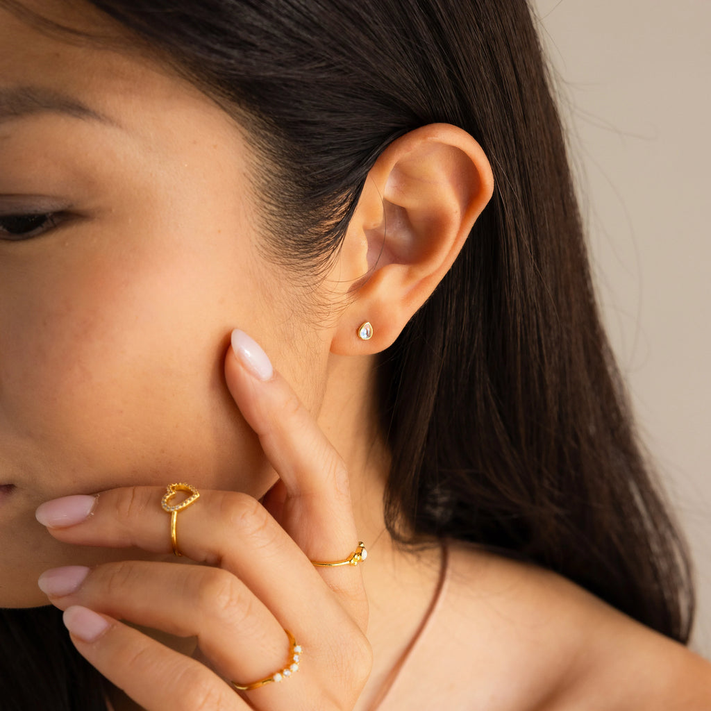 A woman with smooth, long dark hair touches her face, wearing gold rings and the Pear Moonstone Studs for a touch of celestial charm.