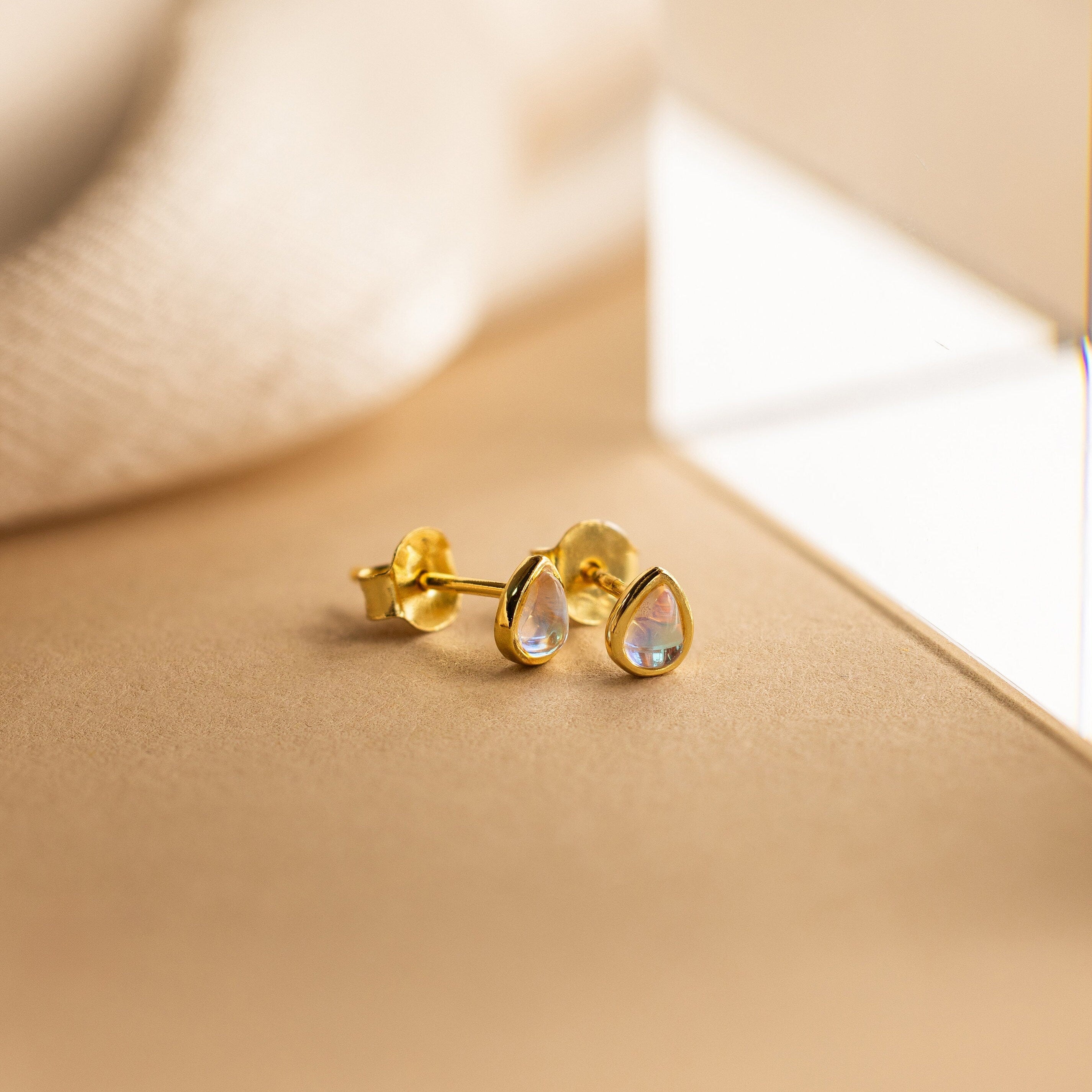 The Pear Moonstone Studs, featuring iridescent stones in a gold teardrop design, radiate celestial charm and elegance against a soft beige background.