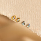 Four dainty Pear Moonstone Studs, featuring teardrop-shaped iridescent stones—two in gold, two in silver—rest on a textured beige surface, radiating celestial charm.