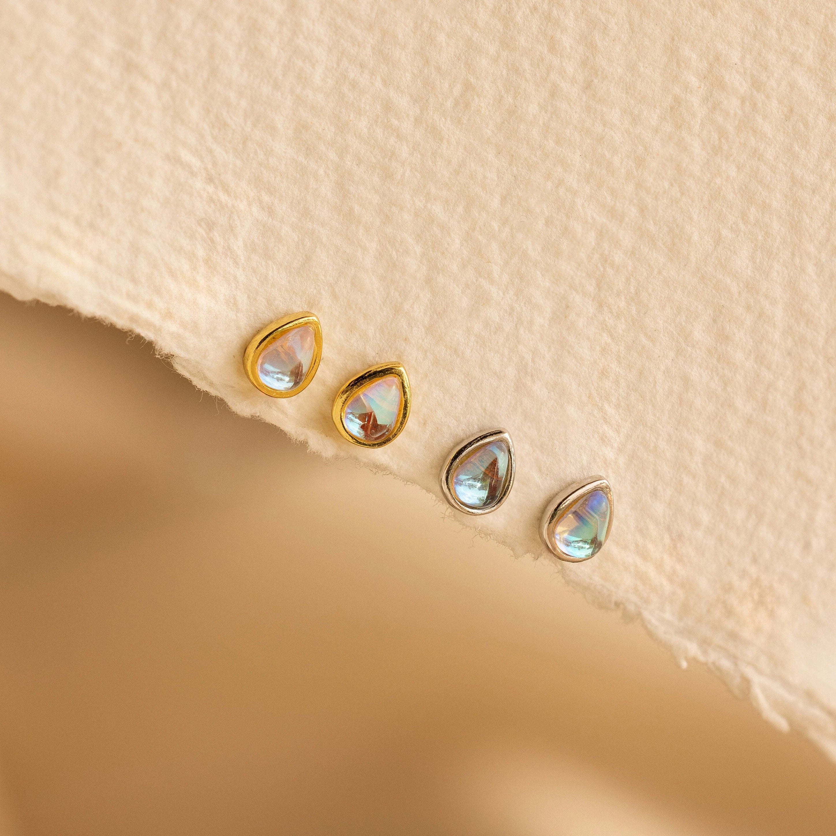 Four dainty Pear Moonstone Studs, featuring teardrop-shaped iridescent stones—two in gold, two in silver—rest on a textured beige surface, radiating celestial charm.