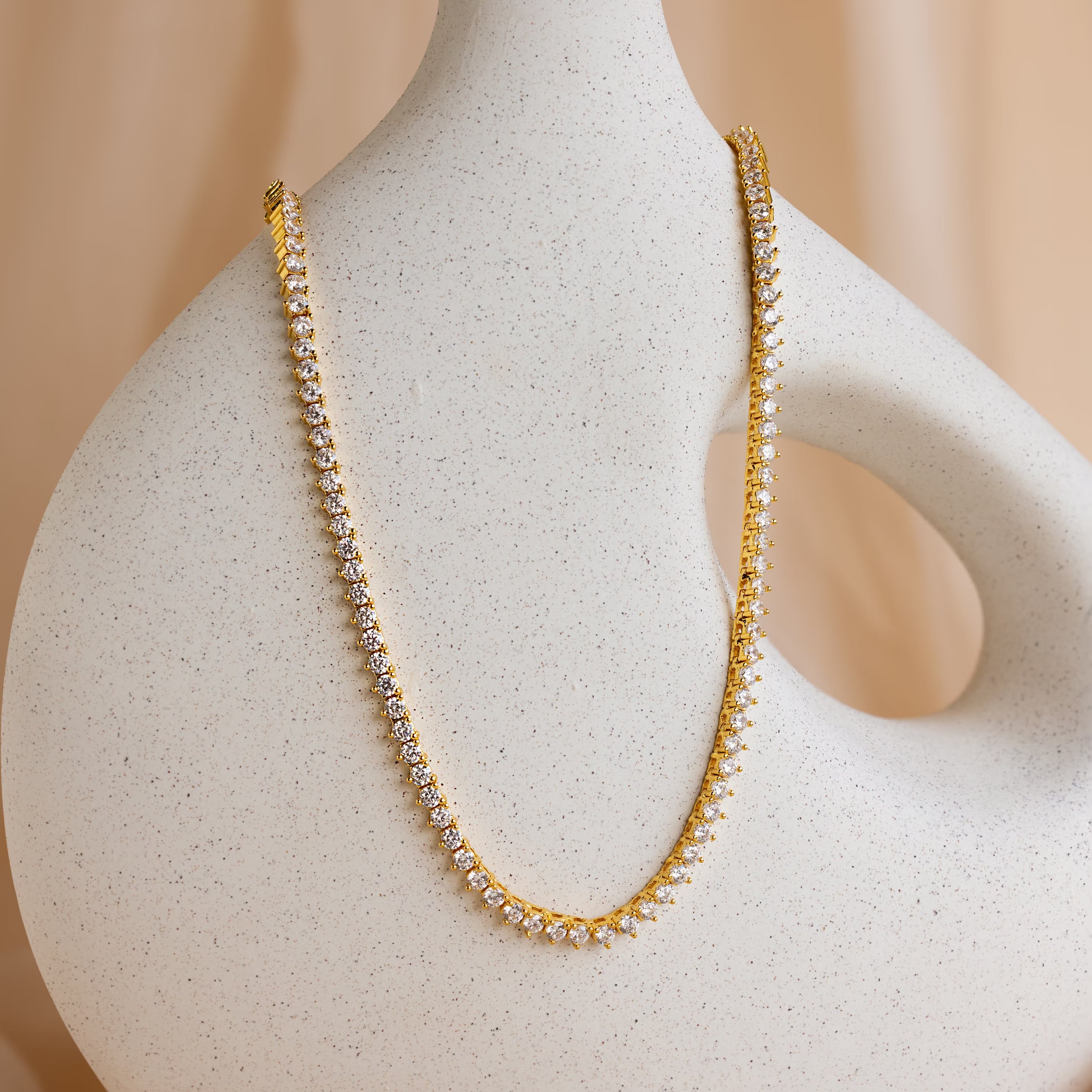The 3 Prong Tennis Necklace is displayed on a white, abstract mannequin with a curved design, highlighting its dainty gold chain and sparkling diamonds.