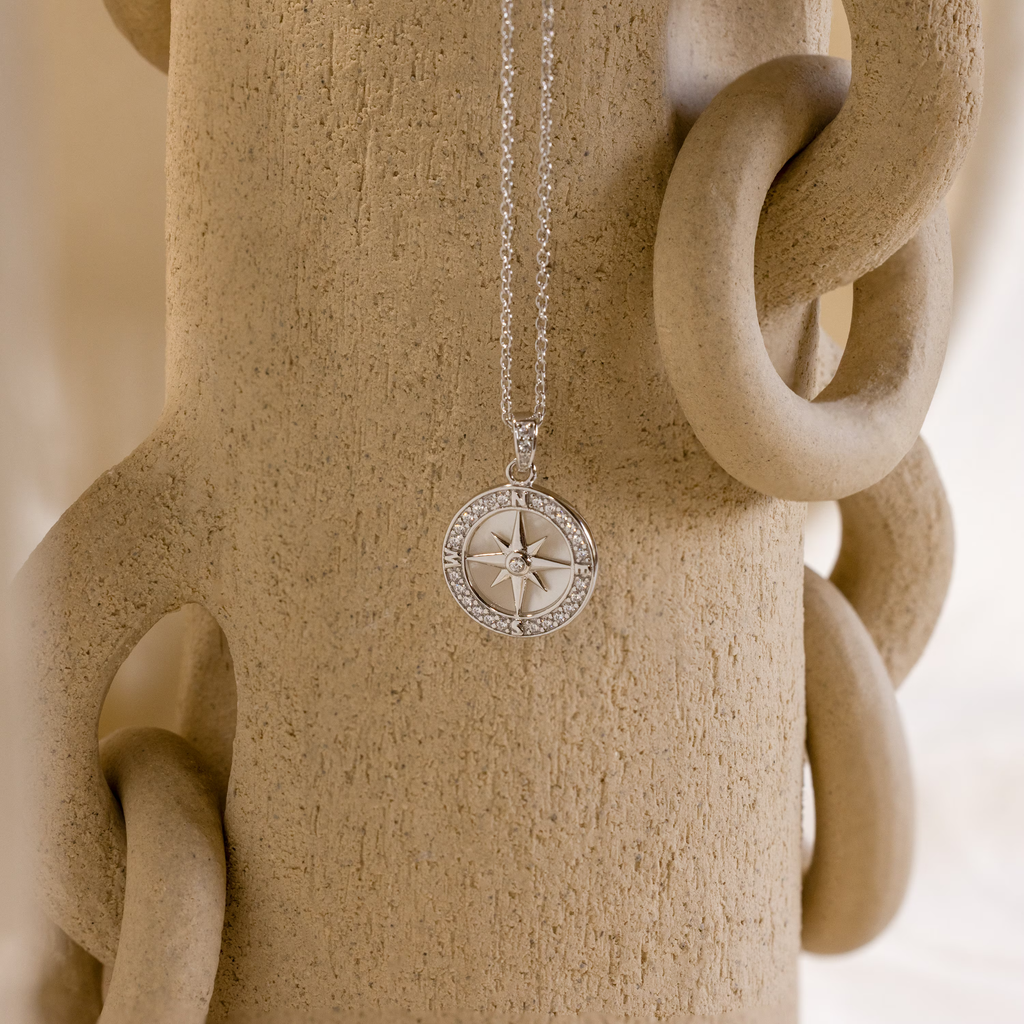 The Pave Pearl Compass Necklace, elegantly displayed on a textured beige vase with circular handles, combines nautical charm with the iridescent beauty of a mother-of-pearl design.