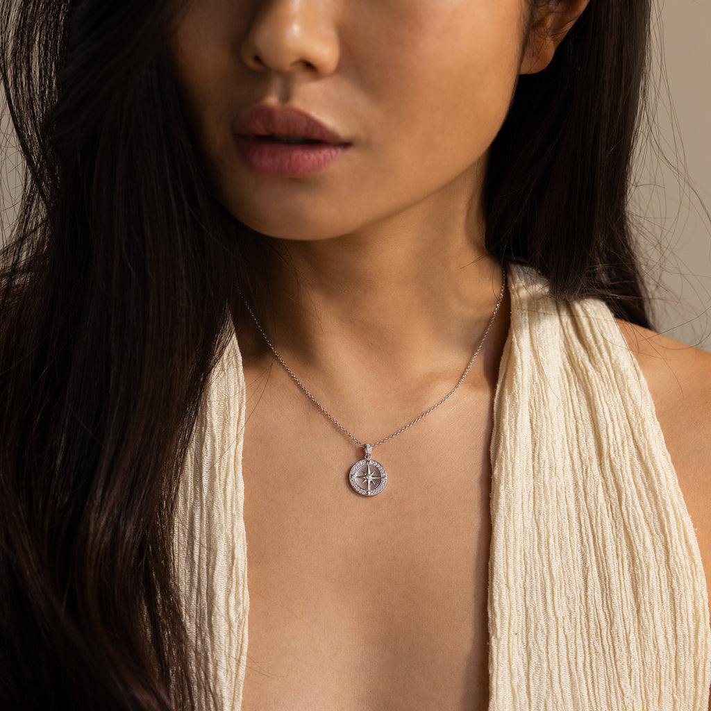 A woman, shown from the shoulders up in a cream textured top, wears the Pave Pearl Compass Necklace—an elegant nautical piece with shimmering details that capture the sea's allure.