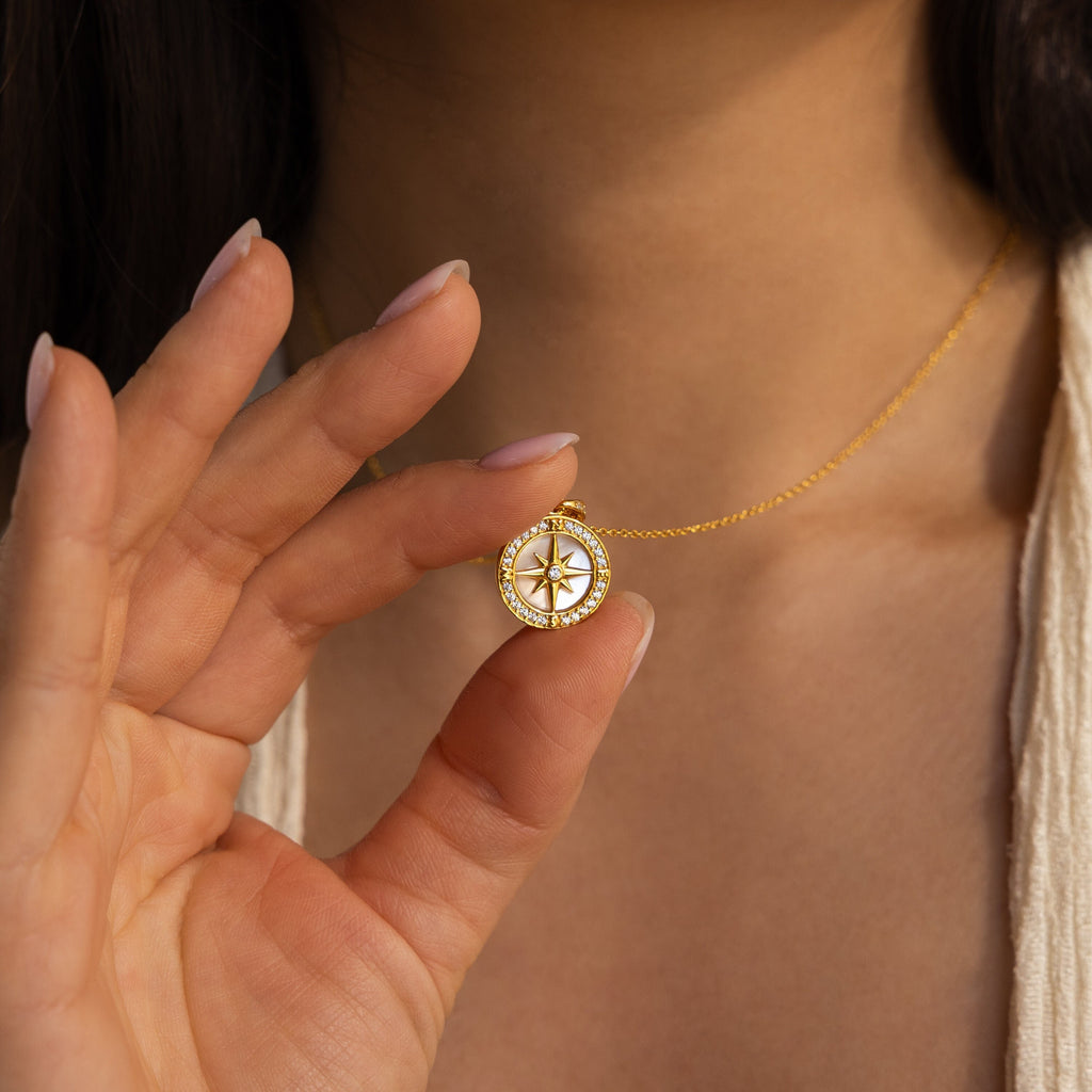 A woman holds the Pave Pearl Compass Necklace near her neck, highlighting the refined elegance of this nautical-inspired jewelry piece.