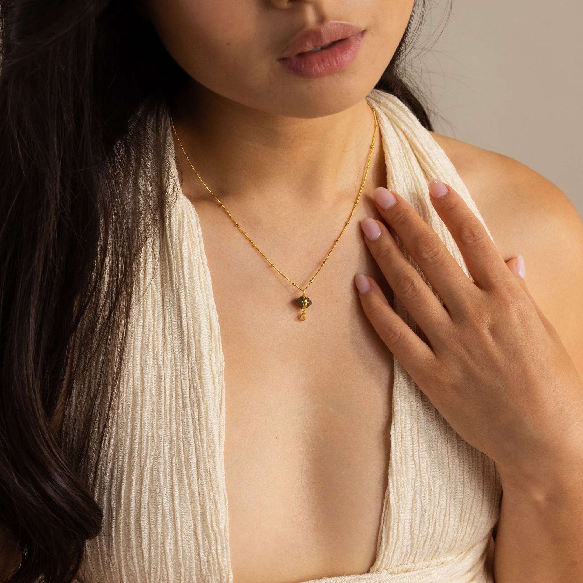 A woman in a cream textured top wears the Labradorite & Moonstone Drop Necklace, a delicate gold drop necklace with a small pendant, as she touches her chest.