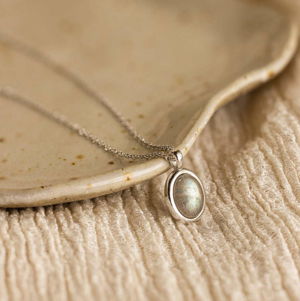 The Oval Labradorite Pendant Necklace features a stunning labradorite gemstone set in a silver oval, beautifully displayed on a ceramic plate with textured fabric underneath.