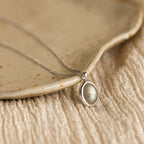 The Oval Labradorite Pendant Necklace features a stunning labradorite gemstone set in a silver oval, beautifully displayed on a ceramic plate with textured fabric underneath.